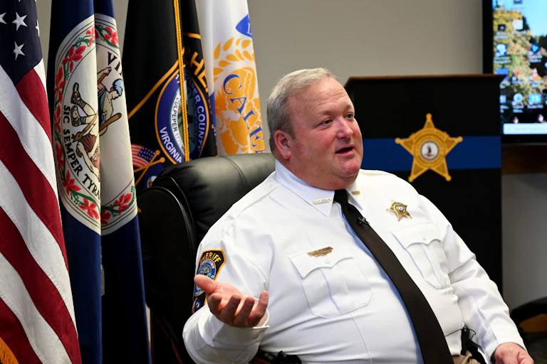 A man in a button-down shirt and tie wearing a badge sits in front of a group of flags.