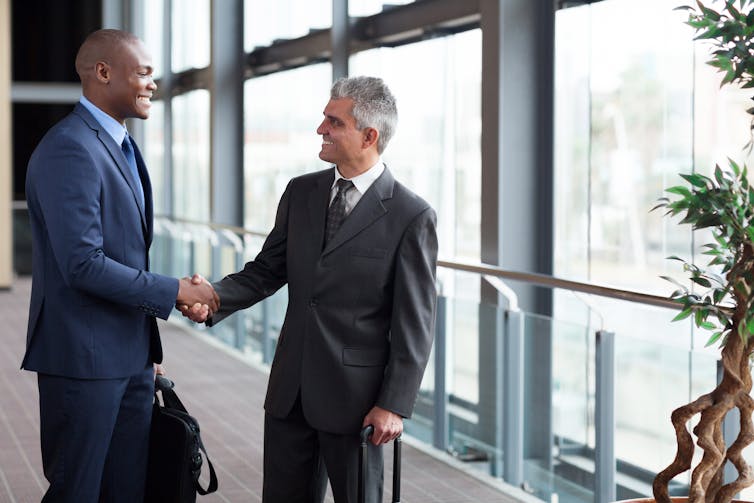Two men shakings hands in front of a row of large windows. One man is holding a briefcase