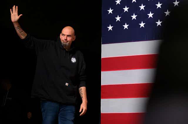 A large, bald man with a goatee and tattoes stands in front of an American flag image.