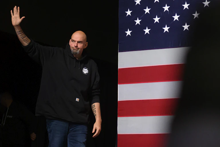A large, bald man with a goatee and tattoes stands in front of an American flag image.