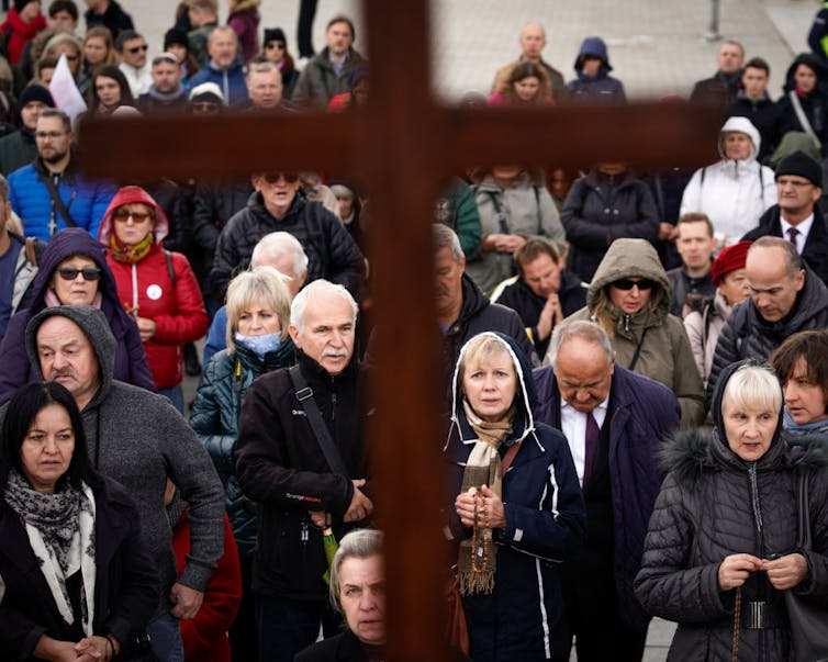 A crowd of people in jackets look up at a tall cross in front of them.