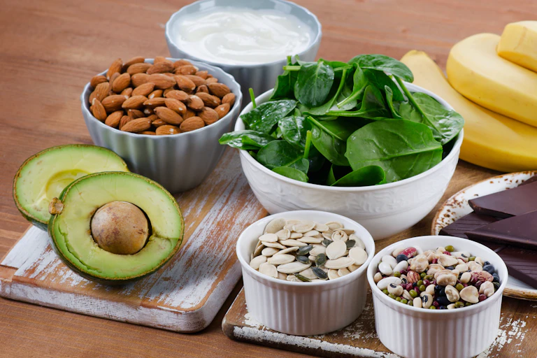 An assortment of magnesium-rich foods on a table, including almonds, spinach, avocado, yoghurt and seeds.