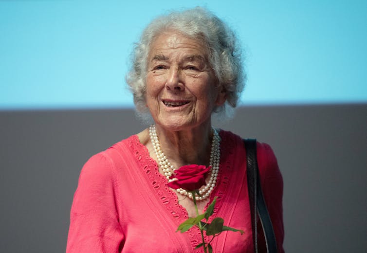 Photograph of a smiling elderly woman wearing pink and holding a red rose