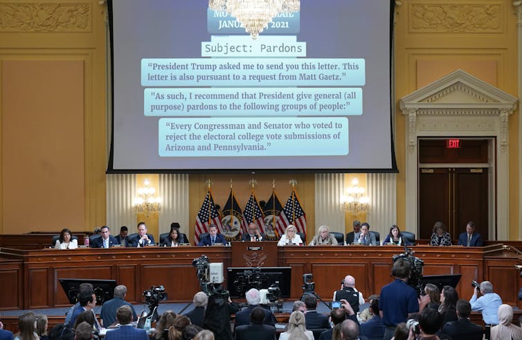 People are seen in a large conference or other official looking room, looking at a large projecter screen that has details about government pardons and Jan. 6.