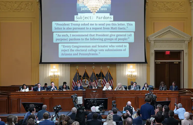 People are seen in a large conference or other official looking room, looking at a large projecter screen that has details about government pardons and Jan. 6.