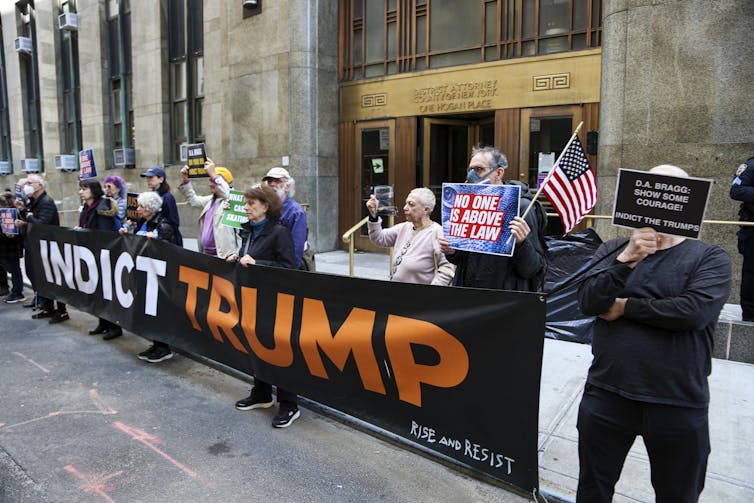 People stand behind a large banner that says 'indict Trump,' and hold up their own small signs in front of an official government building.
