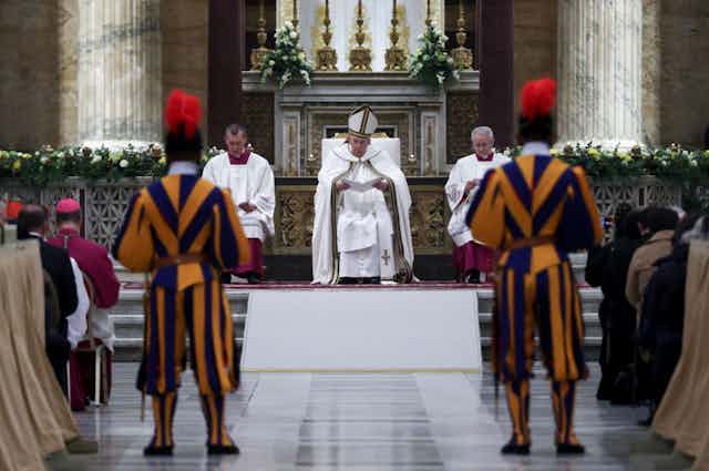 The pope sits on a white chair in a white outfit and tall hat in a cathedral, as two guards in purple and yellow uniforms stand before him.