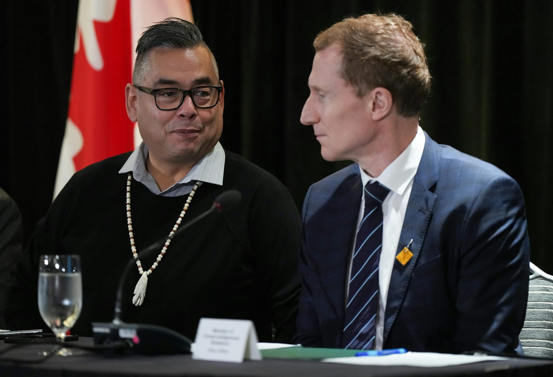 Two men sit at a table. A Canadian flag hangs on a flag pole behind them.