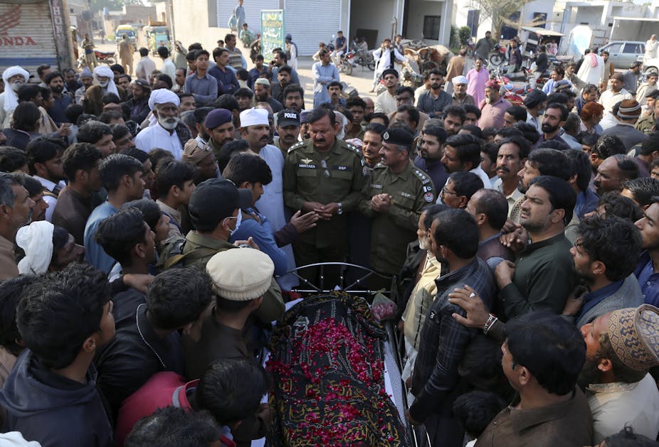A large number of people gathered around an open coffin with a body covered with red flowers.