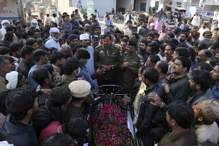 A large number of people gathered around an open coffin with a body covered with red flowers.