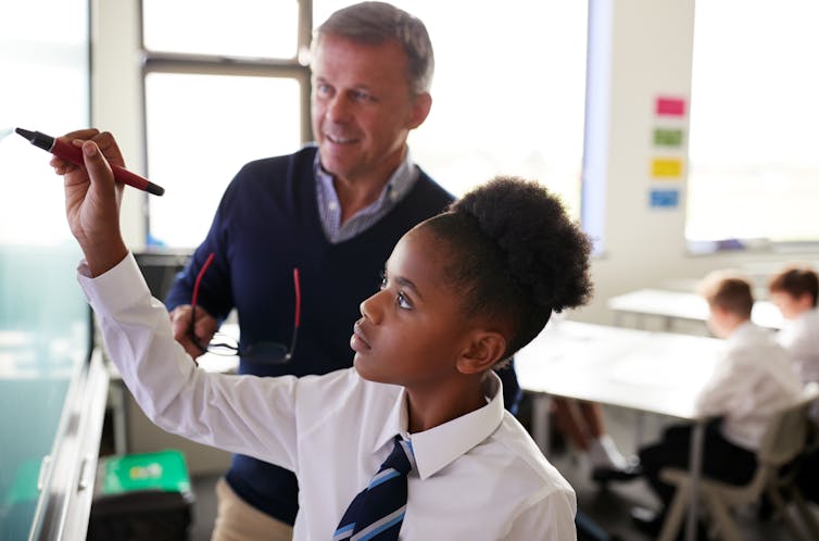 Student writing on a whiteboard with teacher looking on.