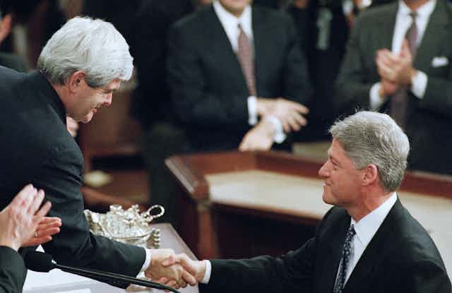 A gray haired white man shakes hands with another gray haired white man, both wearing black suits, surrounding by others wearing black suits