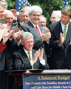 A group of white man clap as president Clinton sits on at desk with the words a balanced budget witten in front