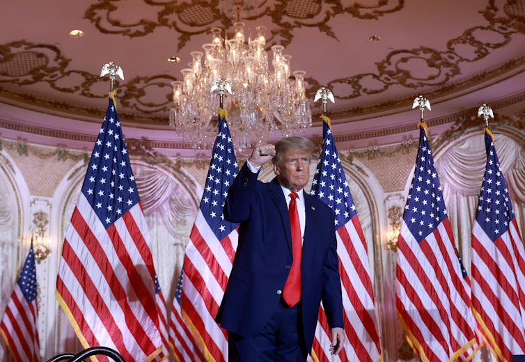 A man in a blue suit, red tie and white shirt showing a clenched fist in front of several US flags.