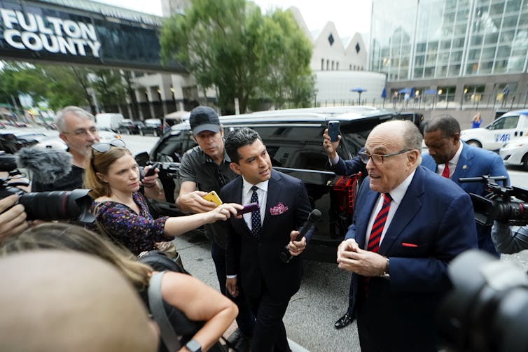 A man in a blue suit, white shirt and red tie, wearing glasses, faces a crowd of reporters with microphones on a sidewalk.