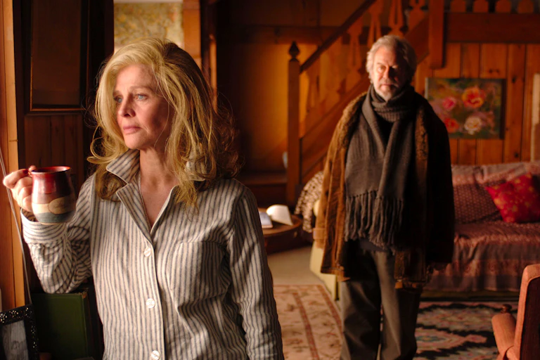 an older man watches a beautiful oder woman, looking out a window, with a mug in hand. Both look sad.