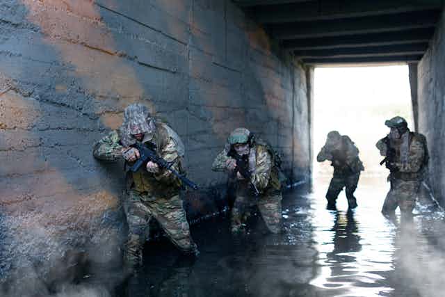Four men in military fatigues wading through a tunnel that has water up to their shins. They are holding guns and are crouched forward.