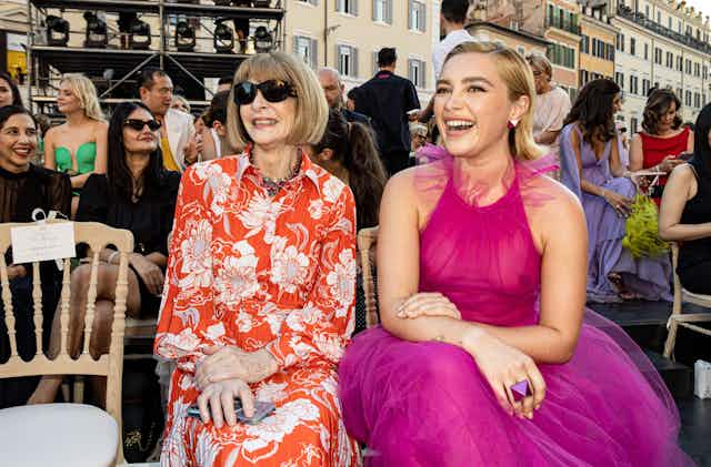 Anna Wintour in sunglasses sits next to a smiling Florence Pugh in a sheer pink dress
