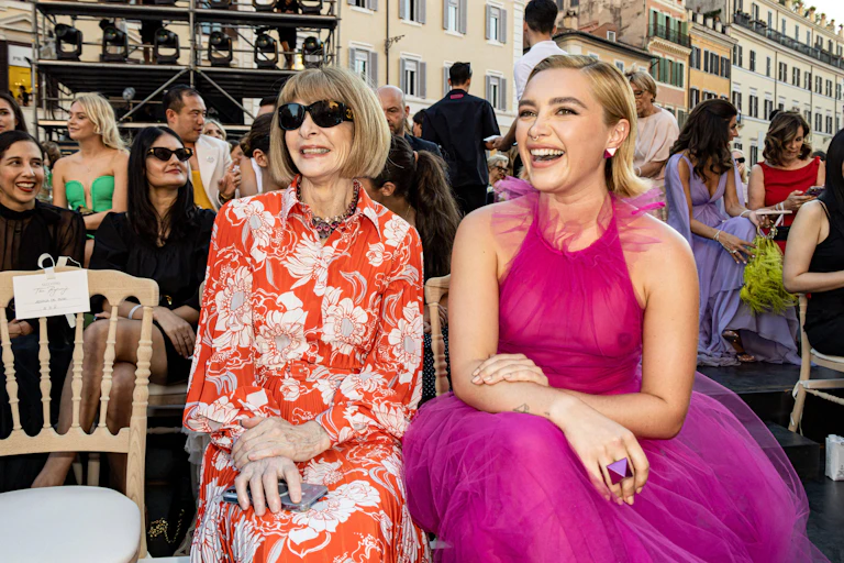 Anna Wintour in sunglasses sits next to a smiling Florence Pugh in a sheer pink dress