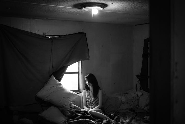 Black and white photo of Alice, a young woman with dark hair, sitting on a bed inside a trailer, with a sheet hanging over the window.