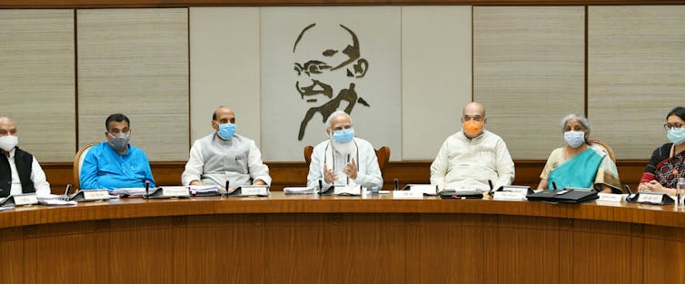Men sitting at a long table wearing medical masks, with an image of Gandhi behind.