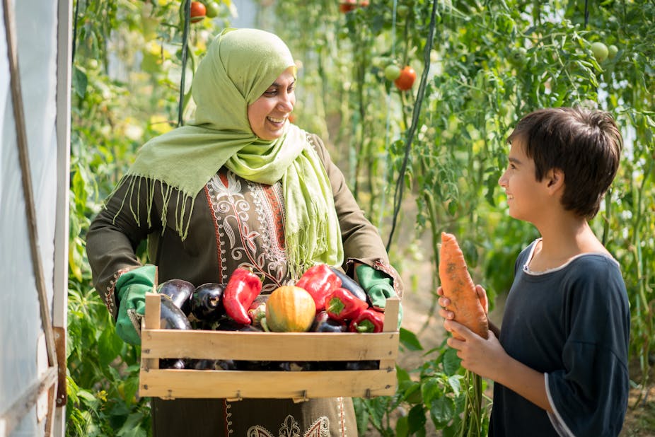 A smiling adult woman wearing green gardening gloves holds a box full of vegetables while a child holding a big carrot chats to her