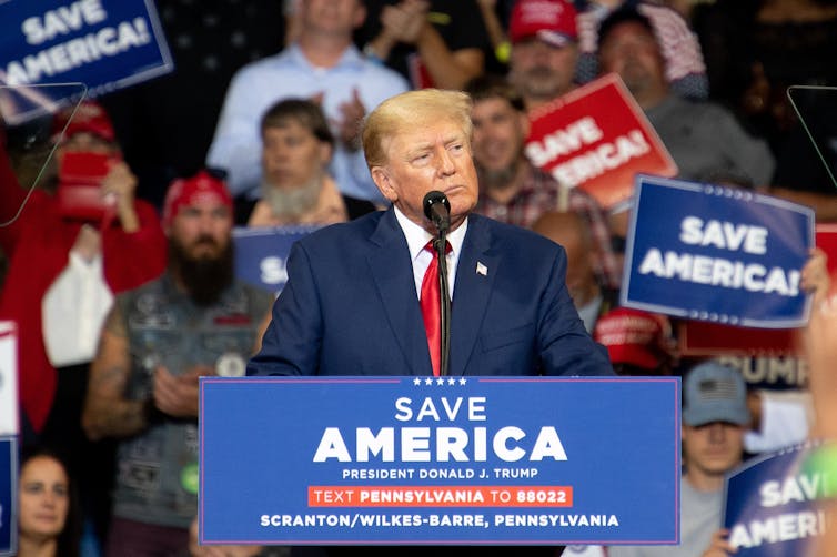 Former US president Donald Trump stands at a lectern in front of a crowd waving 'Save America' placards.gn slogan
