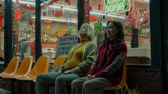 Two women sit on a bench outside of a laundrette. 