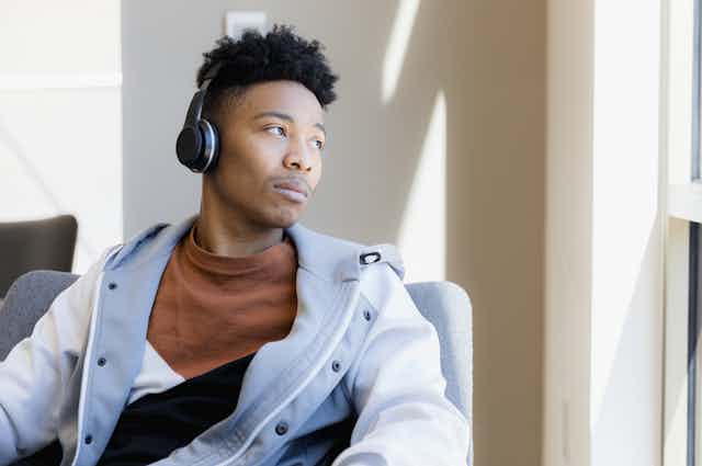 A young Black man wearing headphones looks out the window with a look of resignation.