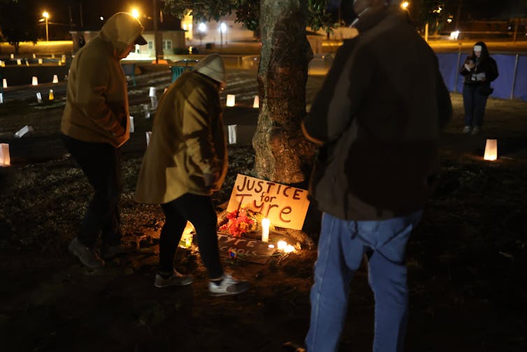 Por la noche, tres personas vestidas con abrigos se paran frente a un cartel que está apoyado contra un árbol e iluminado por velas.  Dice: 