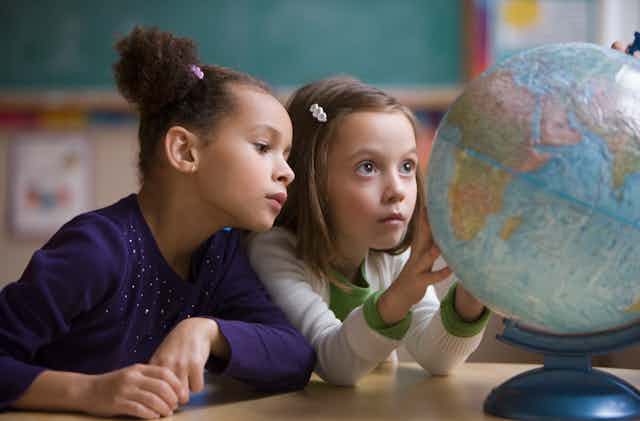 Two young girls look intently at a desktop globe. One has her hands on the globe as if she's rotating it.