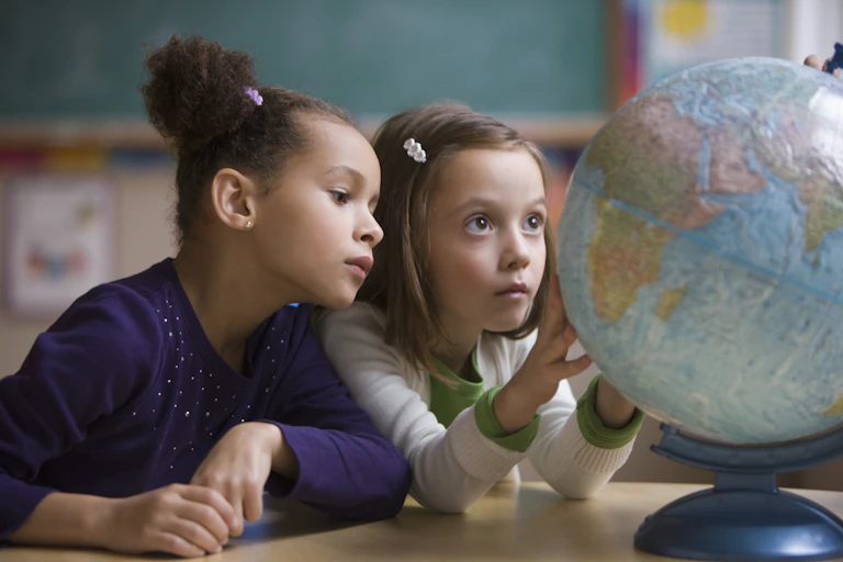 Two young girls look intently at a desktop globe. One has her hands on the globe as if she's rotating it.