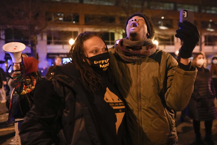 Demonstrators gather on a street, one crying out and the other with a face mask with 'defund MPD' written on it.