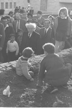 A man with tufts of white hair, wearing a suit, watches children plant a small tree in a black and white photo.