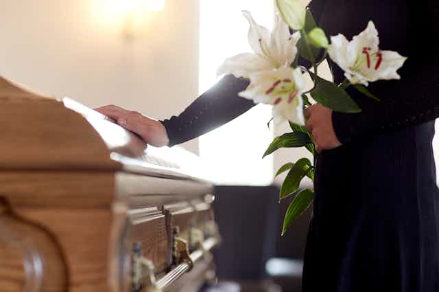 A person gently touches a coffin while holding white flowers.