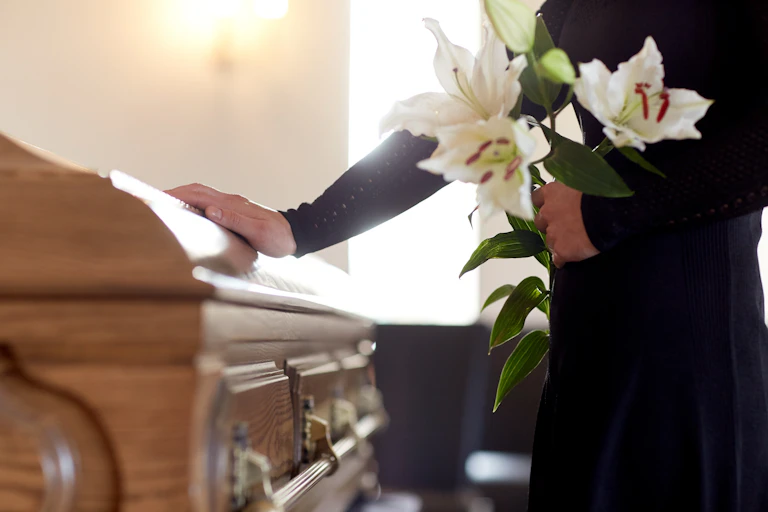 A person gently touches a coffin while holding white flowers.