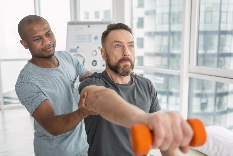 A man performs an exercise using a dumbbell while his trainer helps him.
