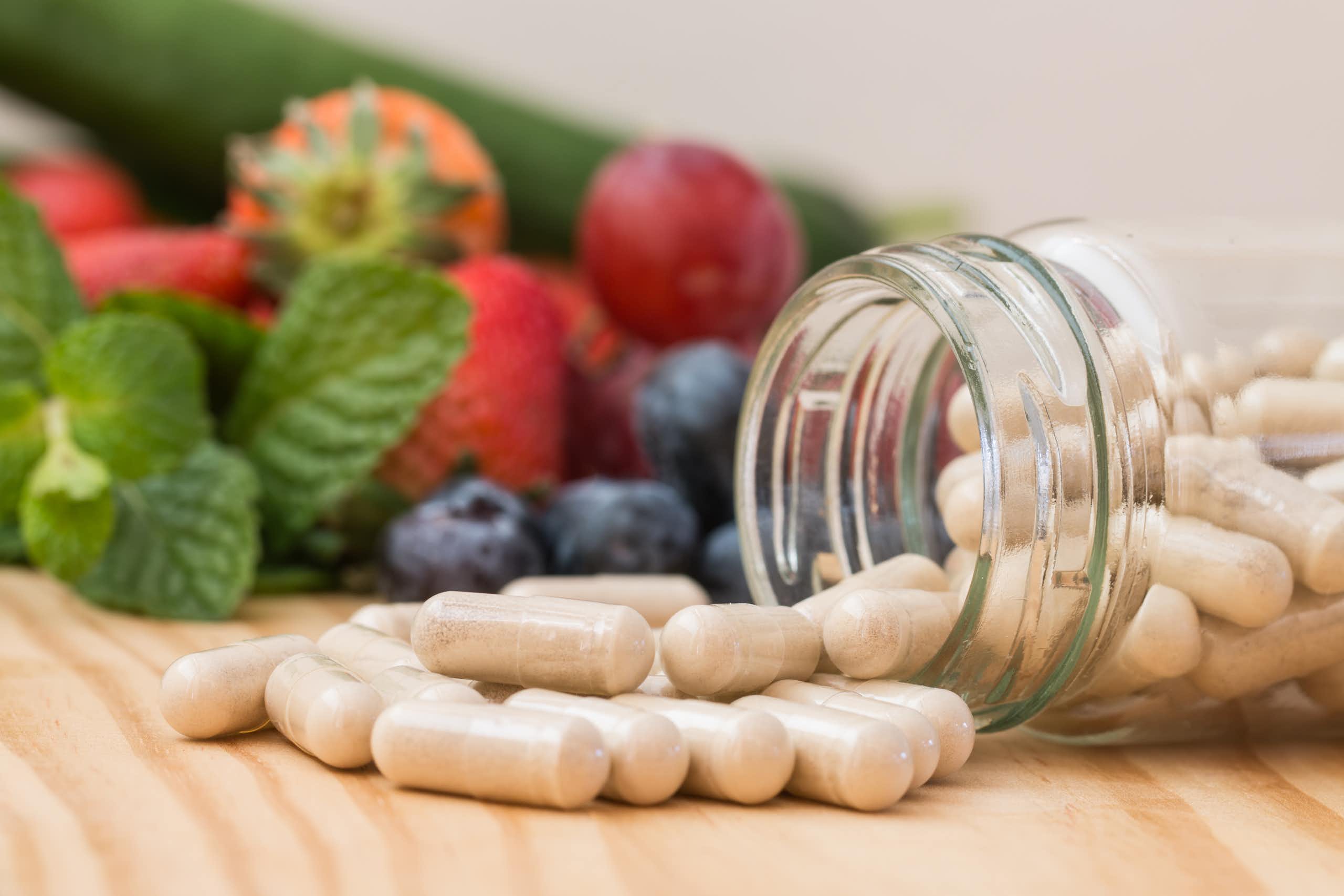 Beige pills pouring out of in bottle on a wooden table, with blueberries, strawberries and grapes in the background.