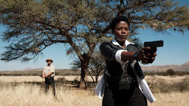 In an arid landscape under a large tree a Namibian woman points a gun straight ahead of her. Behind her a man holds a lowered gun.