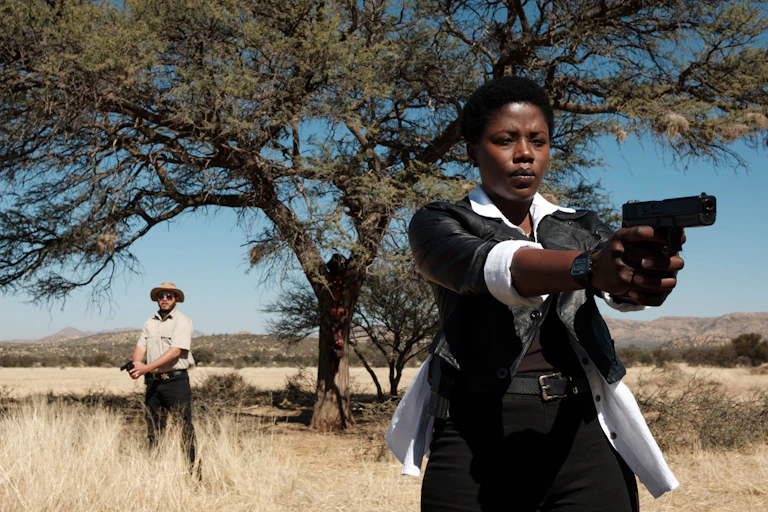 In an arid landscape under a large tree a Namibian woman points a gun straight ahead of her. Behind her a man holds a lowered gun.