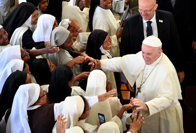 An elderly man in Catholic pope's outfit smiles as he reaches out both hands to touch the outstretched hands of a group of smiling African women in nun's habits.