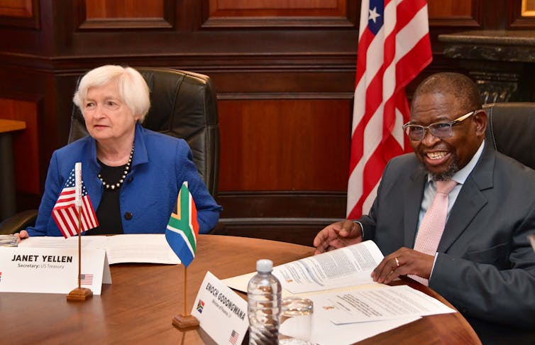A man and a woman smile for the camera while sitting. Miniature South African and America flags are on the table.