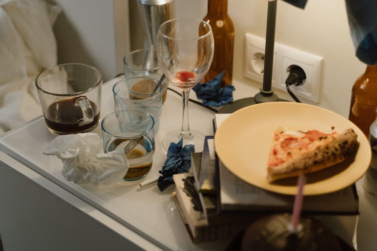 A messy side table with a beer bottle, a wine glass, several other glasses and a plate with a slice of pizza on it