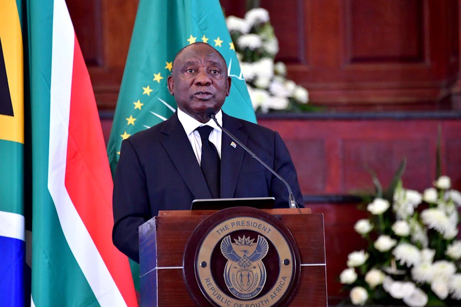 A man wearing a suit and tie speaks at a podium bearing the insignia: 'Presidency of South Africa, Republic of South Africa' with his back to the South African and African Union flags. 