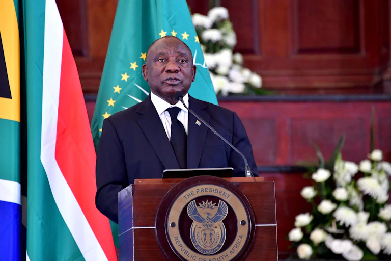 A man wearing a suit and tie speaks at a podium bearing the insignia: 'Presidency of South Africa, Republic of South Africa' with his back to the South African and African Union flags.