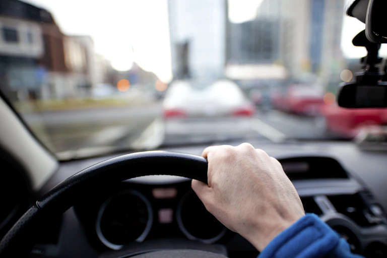 Right hand is seen on wheel of car with other cars in front outside the windshield