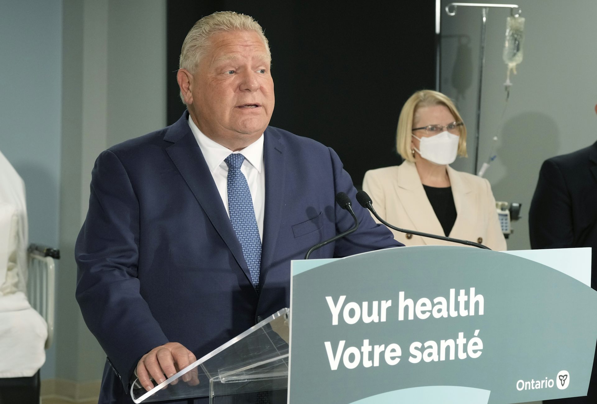 A man in a dark suit speaking at a podium with a sign that reads: Your heath, Votre sante.