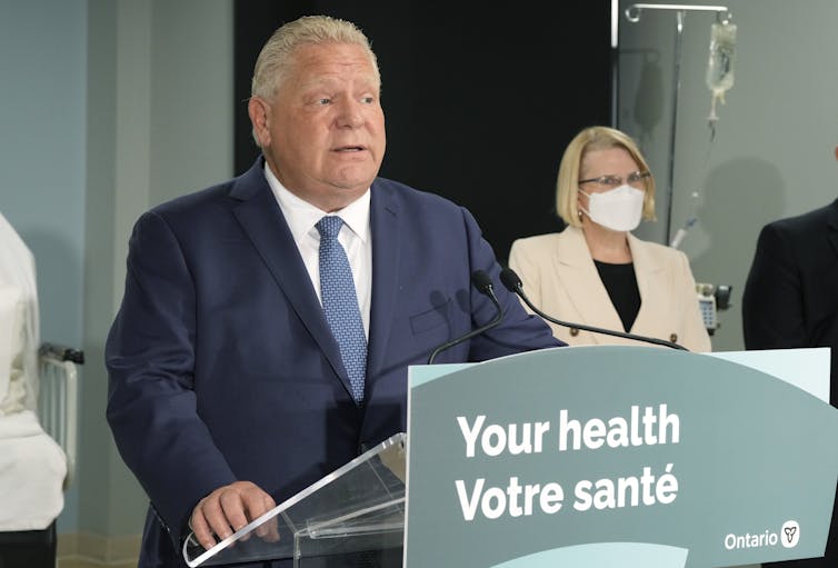 A man in a dark suit speaking at a podium with a sign that reads: Your heath, Votre sante.