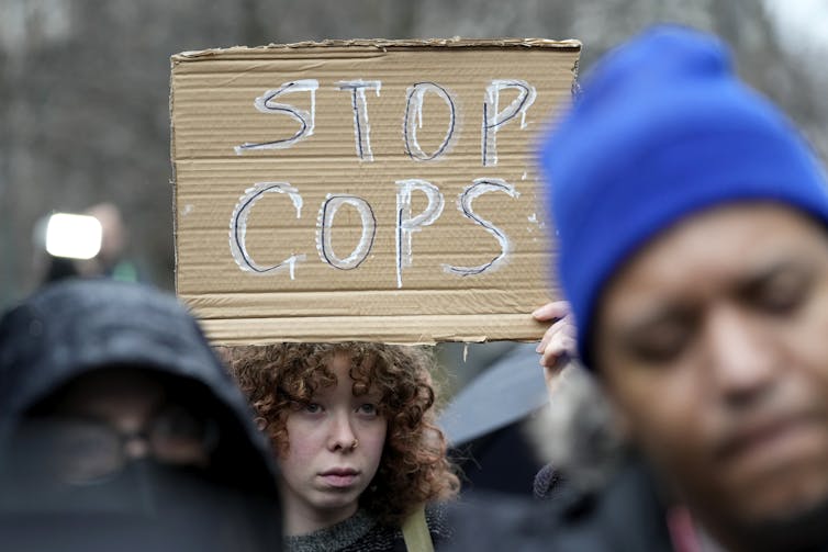 A woman holds up a sign that says Stop Cops. Two Black people are in the foreground.