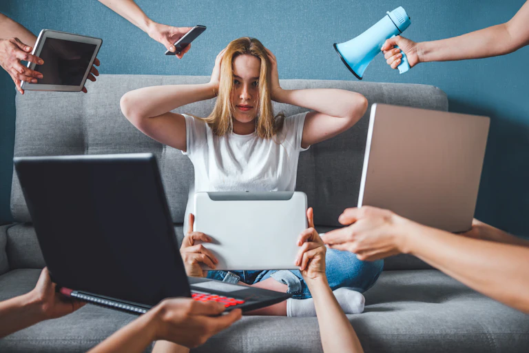 A woman sits on a sofa covering her ears while screens are shown facing her from multiple directions.
